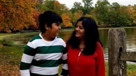 A teenager of Indian heritage  with a green and white striped shirt and looks at his mother standing in a red shirt in front of colorful fall trees 