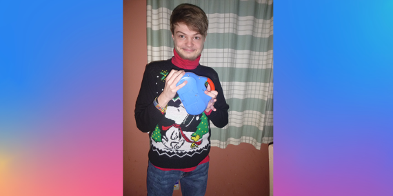 A young man stands in a black sweater with a chistmas tree graphic with Snoopy and Woodstock on it. With a red shirt underneath. He is smiling at the camera while holding a toy clock radio 