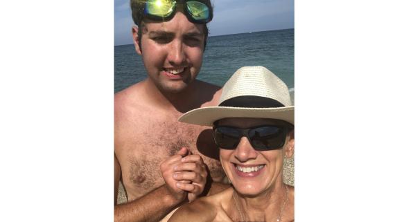 A mother and son are standing in front of the ocean for a selfie. The son has green googles on the top of his head, his mom has a white straw hat and is smiling. They both are smiling