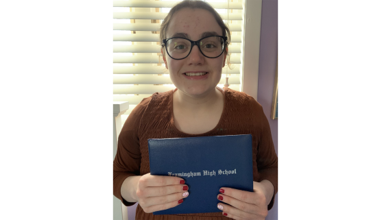 A girl with black glasses and a maroon shirt holds a blue high school diploma to the camera and is smiling 