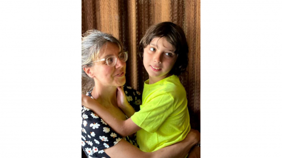 A young boy in a  bright yellow shirt looks out in the distance in his mother's arms, as the mother in glasses and a polka dot shirt looks at him 