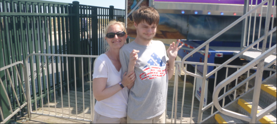 A mother stands behind her teenage son at a outdoor broadwalk setting. She has blonde hair and sunglasses and he is wearing a grey t-shirt with an American flag logo on it 