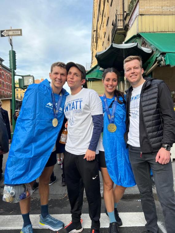 A man and a women in post marathon blue coats wear gold medals and smile with two other men in shirts that read Team Wyatt 