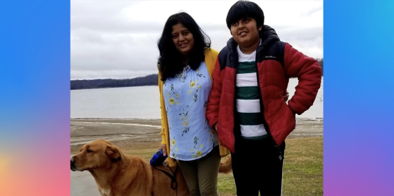 A yellow lab dog stands beside a mother and son who are smiling. The mother is in a yellow  sweater and a blue top and the son has his hands on his hips and has a red puffer jacket on. They are standing in front of a lake on a cloudy day 