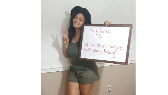 A young women wearing green and a black hat, makes a peace sign with one hand as she holds a whiteboard sign that says "Victoria, 28: Stim: stick tongue out when thinking" 
