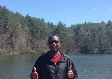 Ryan Lee wearing sun glasses and standing in front of a lake surrounded by trees