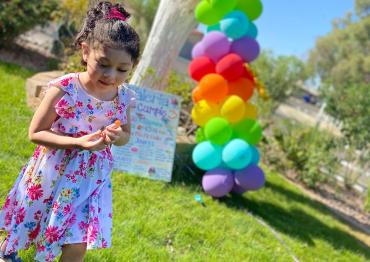 A child in a dress standing in the grass next to a tree with balloons