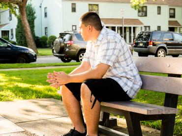 A young man wearing a plaid shirt while sitting on a bench outside and looking away at the road