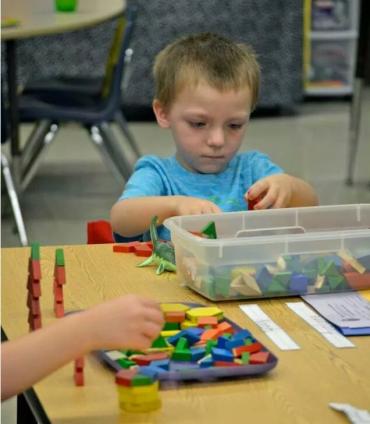 young boy sorting blocks at school desk with plastic bin of building blocks in front of him