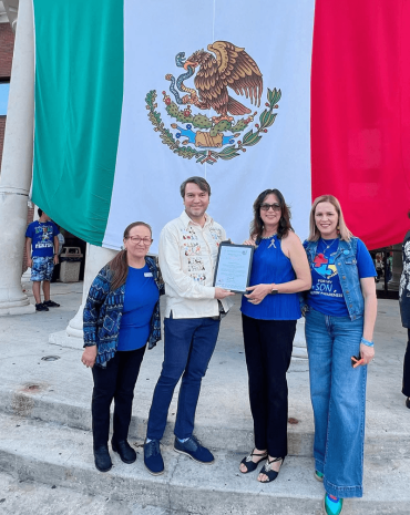 Four people pose in front of a large Mexican flag, with one man and a woman holding a framed certificate