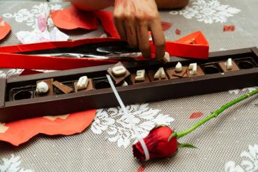 Hands arranging I LOVE YOU chocolate letters in a box next to a red rose