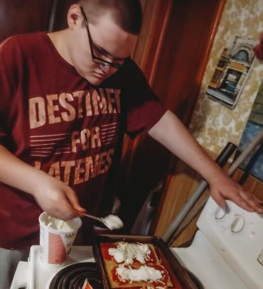 A man in a red t-shirt stands over a white stove and places cheese on pasta sheets