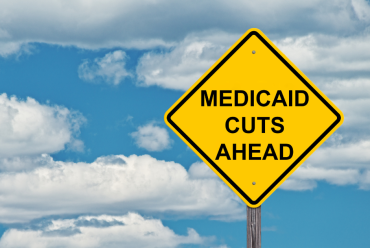 Yellow road sign that says "MEDICAID CUTS AHEAD" with blue sky and clouds in background