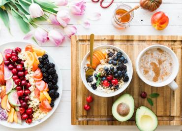 Overhead view of breakfast spread with yogurt bowl, fresh fruit, avocado halves, coffee, honey jar, and tulips on white table