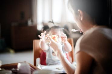 a toddler in a highchair reaching for food as her mother feeds her
