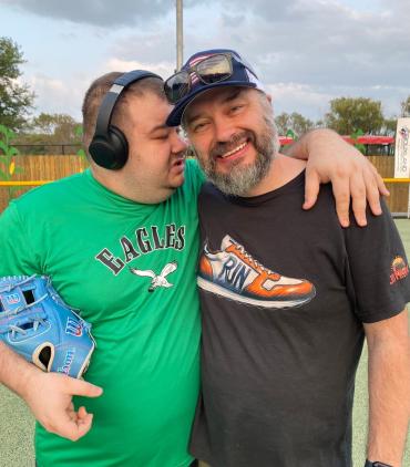 man in green eagles shirt holding baseball and hugging woman on a green turf field at sunset