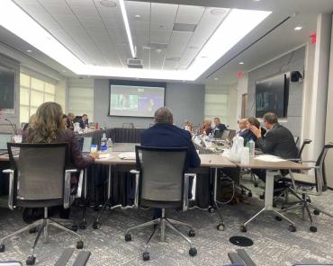 rectangular shaped conference table with people sitting around it and projector with video conference playing on farthest wall