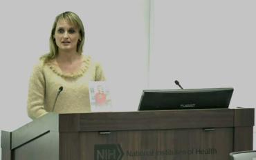 woman in yellow blouse standing behind large wooden podium with microphone speaking