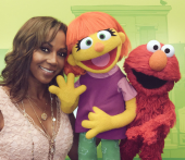 A woman smiling next to Elmo and Julia from Sesame Street