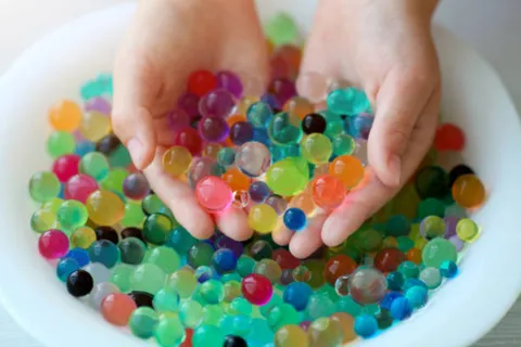 two hands holding a bunch of colorful water beads over a white bowl of more water beads